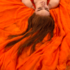 Rainbow Squared, Year 5, Piece Forty-Eight: 9. Orange Orange. An animated loop using as background a still image of a white-skinned red haired person lying on an orange piece of flowy fabric, head visible from the eyes down upside down in the upper right corner. Her long hair is flowing down, draped on the fabric. Glowing orange lines animate as strands of hair, finally all illuminated at once. Her eyebrows begin to illuminate hair by hair as the background switches to psychedelic swirls, then disappearing.